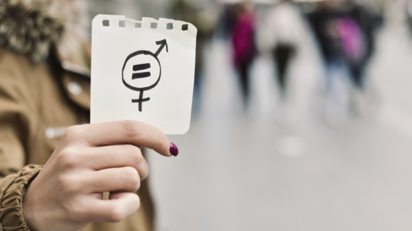 Auf dem Bild ist der Oberkörper einer Frau zu sehen, die einen Notizzettel in das Bild hält. Darauf zu sehen sind die verflochtenen Piktogramme für Männer und Frauen und darin ein =-Zeichen.